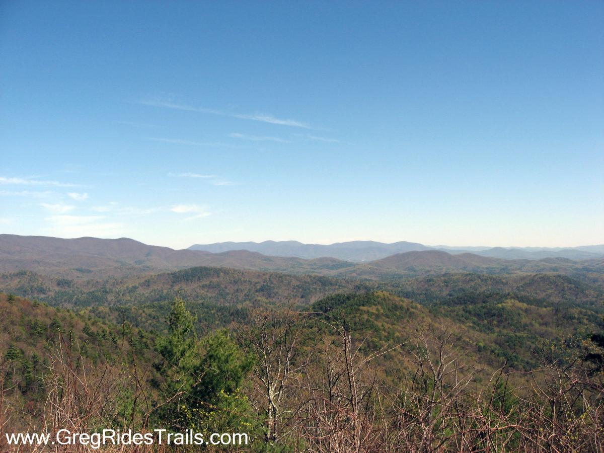 A panoramic view of rolling mountains under a clear blue sky, showcasing various shades of green and brown foliage on the slopes. The landscape features a mixture of trees and hills in the foreground, with more distant mountain ranges fading into the horizon. Bear Creek mountain bike trail.