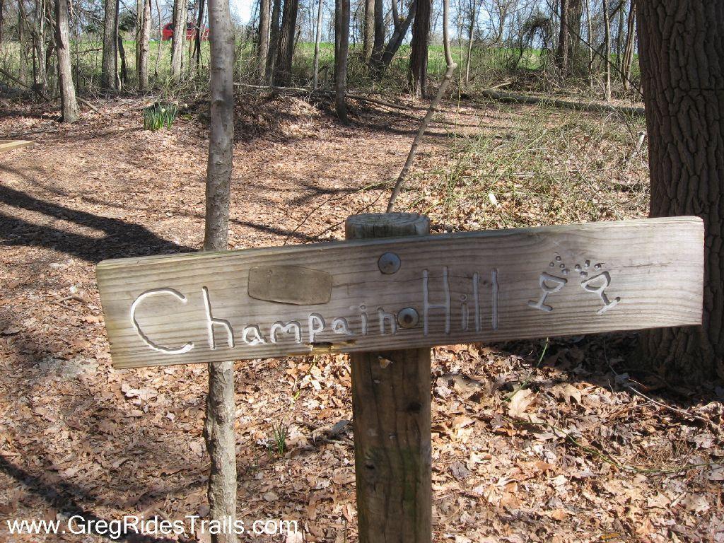 Wooden trail sign labeled "Champaino Hill" with playful illustrations of cocktail glasses, surrounded by a wooded area with fallen leaves and trees in the background. Chicopee Woods mountain bike trail.