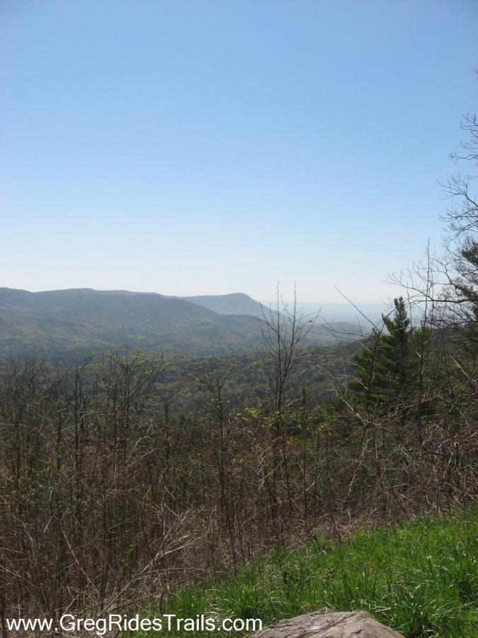 A panoramic view of rolling hills and mountains under a clear blue sky, with sparse trees and greenery in the foreground. The landscape features a mix of distant peaks and valleys, suggesting a peaceful outdoor setting. Bear Creek mountain bike trail.