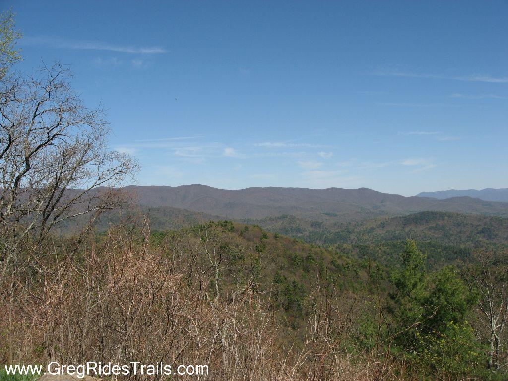 Scenic view of rolling mountains under a clear blue sky, featuring a mix of bare trees and greenery in the foreground. The landscape showcases layers of mountains extending into the distance, creating a peaceful and natural atmosphere. Bear Creek mountain bike trail.