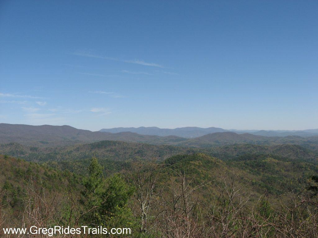 A panoramic view of rolling hills and mountains under a clear blue sky, featuring a mixture of green forests and muted earth tones, showcasing the natural beauty of the landscape. Bear Creek mountain bike trail.