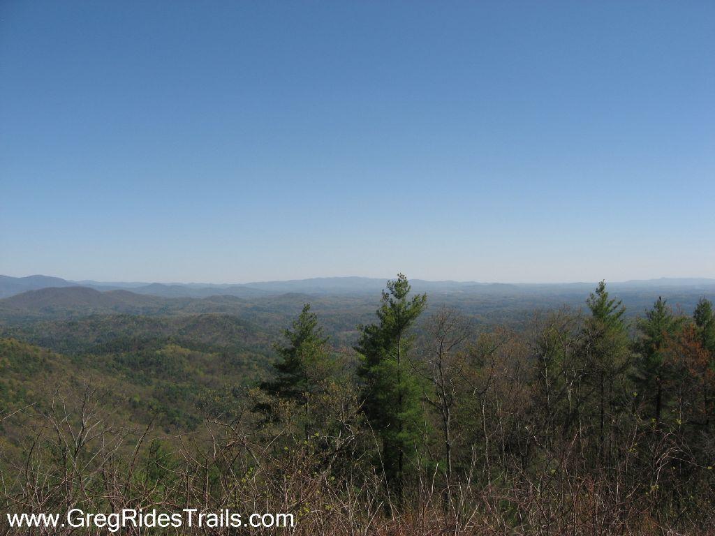 A panoramic view of rolling hills and valleys under a clear blue sky, with a foreground of evergreen trees. The landscape features a mix of lush green and brown foliage, suggesting a transition between seasons. Bear Creek mountain bike trail.