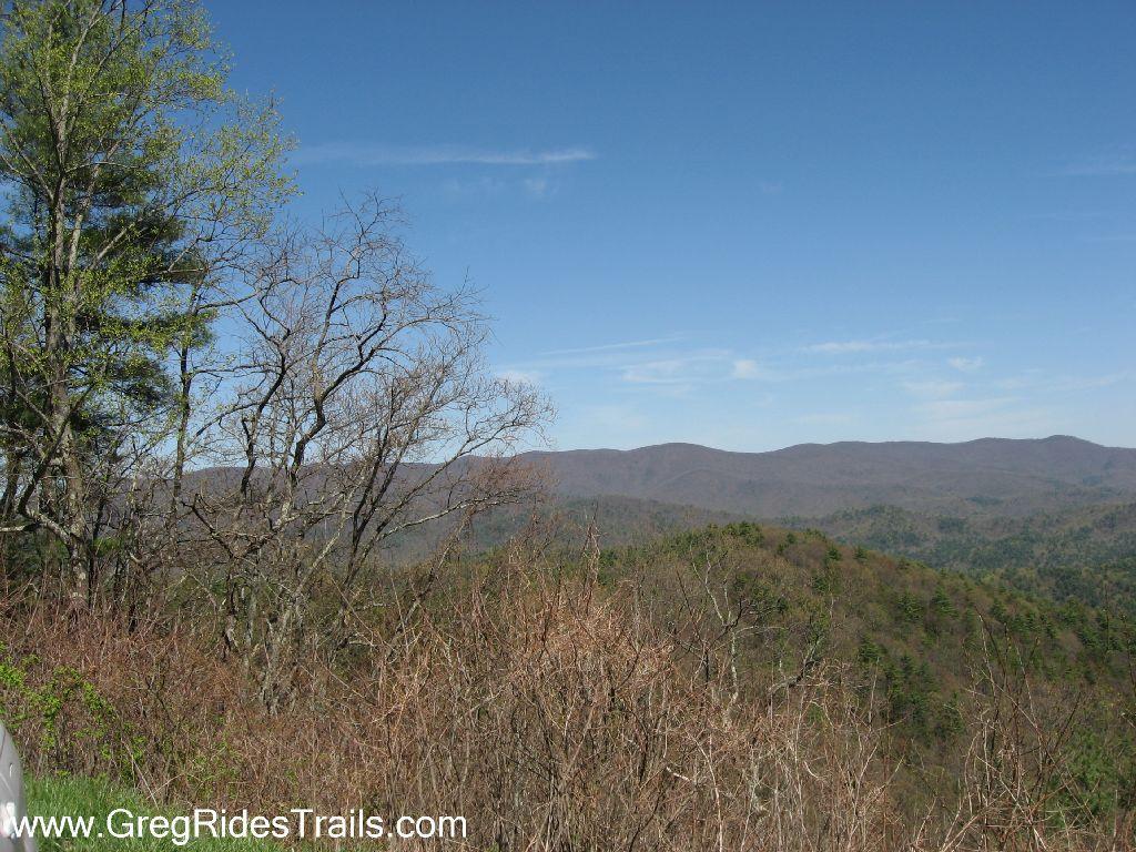 A panoramic view of rolling mountains under a clear blue sky, featuring a mix of green and bare trees in the foreground. The landscape showcases layers of mountain ridges in the background, displaying shades of green and brown. Bear Creek mountain bike trail.