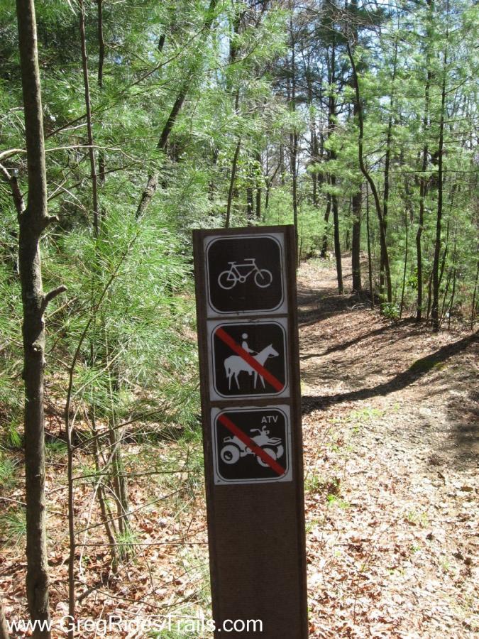 A trail sign surrounded by trees, featuring symbols indicating allowed and prohibited activities: a bicycle symbol indicating biking is allowed, a "no" symbol for horseback riding, and a "no" symbol for ATVs. The trail appears natural with a dirt path and scattered leaves on the ground. Bear Creek mountain bike trail.