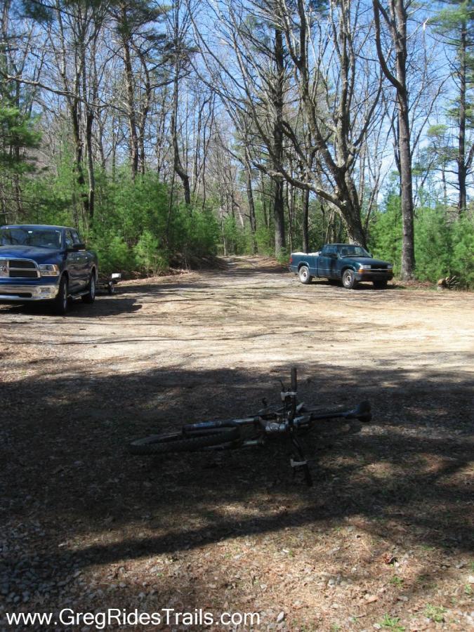 A gravel road through a wooded area, with two parked pickup trucks on either side and a mountain bike lying on the ground in the foreground. The scene is sunny, with leafless trees and some evergreen bushes in the background. Bear Creek mountain bike trail.