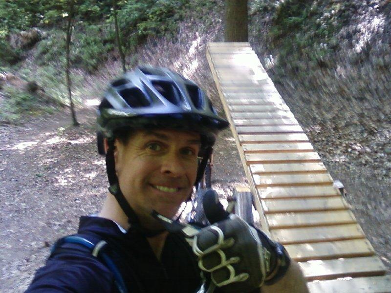 A person wearing a helmet and gloves smiles while giving a thumbs-up in front of a wooden bike ramp in a forested area. Angler's Ridge mountain bike trail.