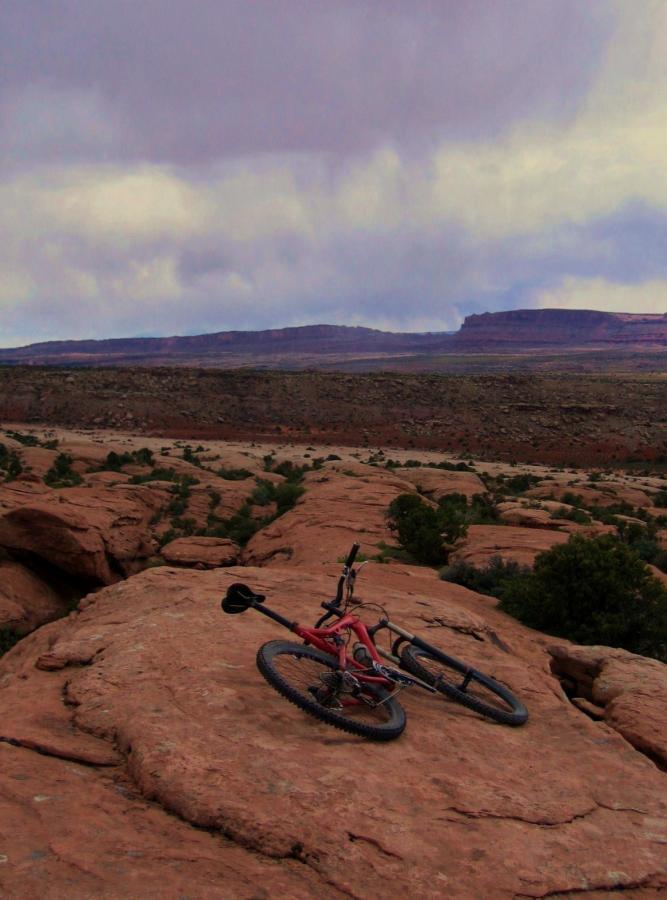 A mountain bike resting on a rocky surface, overlooking a vast, rugged landscape under a cloudy sky. The terrain features red rock formations and patches of greenery in the distance. Sovereign Single Track mountain bike trail.
