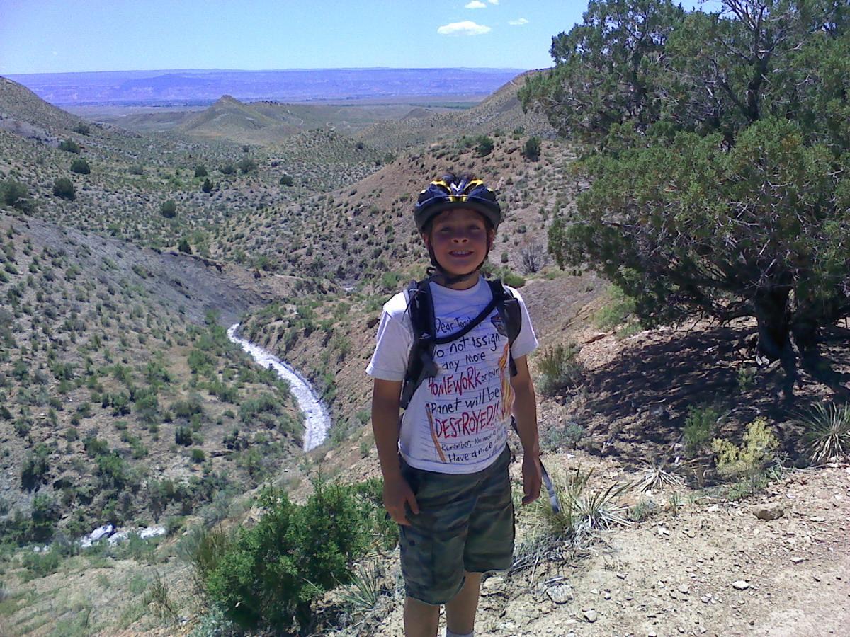 A young boy wearing a bicycle helmet and a white T-shirt with a handwritten message stands on a hiking trail. He is smiling and surrounded by a scenic landscape of hills and greenery, with a winding stream visible in the valley below. The sky is clear with a few clouds, suggesting a sunny day. Chutes and Ladders mountain bike trail.