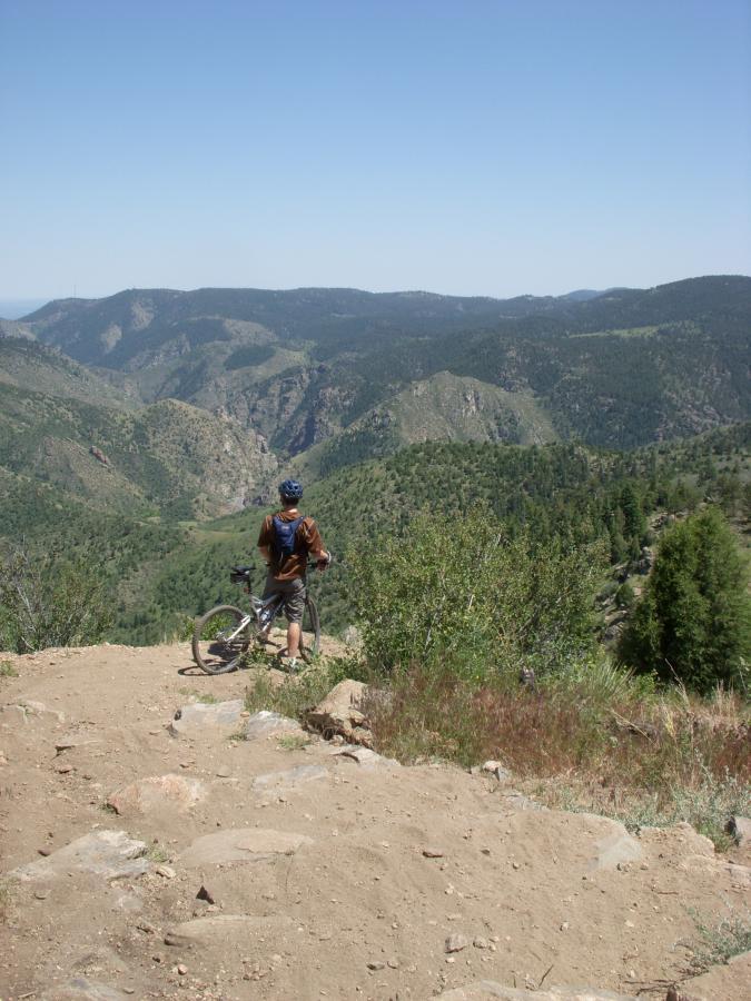A person standing next to a mountain bike, looking out over a scenic view of rolling mountains and lush green valleys under a clear blue sky. Centennial Cone Park mountain bike trail.