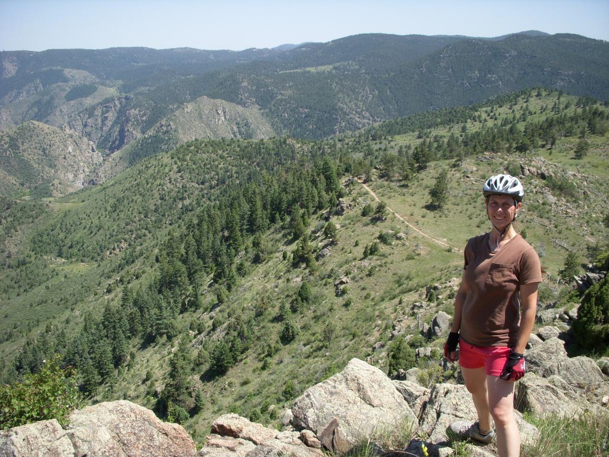 A person wearing a bike helmet and gloves stands on a rocky outcrop, smiling at the camera. In the background, a panoramic view of rolling green hills and dense forests stretches into the distance under a clear blue sky. A trail is visible winding through the landscape. Centennial Cone Park mountain bike trail.