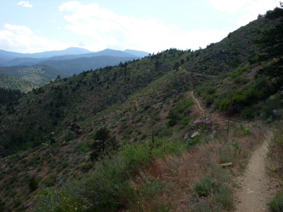 A winding dirt trail through a hilly landscape, surrounded by sparse vegetation and rocky terrain. The background features rolling mountains under a partly cloudy sky. Centennial Cone Park mountain bike trail.