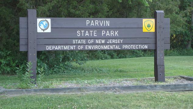 A wooden sign marking the entrance to Parvin State Park in New Jersey, displaying the park's name, the state name, and the Department of Environmental Protection emblem. The sign is surrounded by green grass and trees. Parvin State Park mountain bike trail.