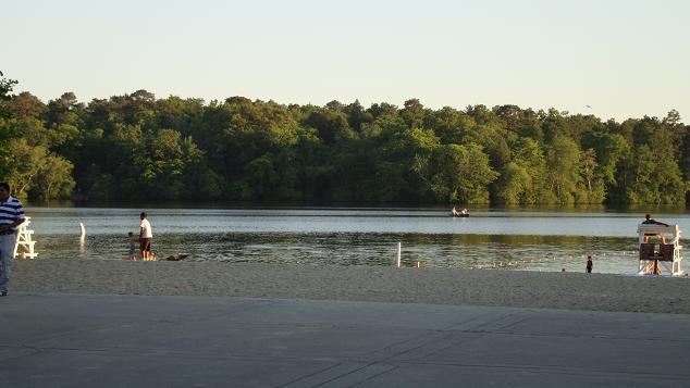 A peaceful beach scene at sunset, featuring a calm lake surrounded by lush greenery. People are enjoying the water, with a sand area in the foreground and a lifeguard station visible. Trees line the background, creating a serene atmosphere for relaxation and leisure activities. Parvin State Park mountain bike trail.