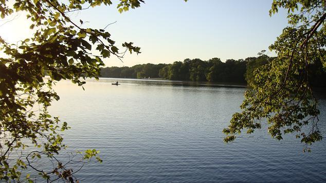 A serene view of a calm lake surrounded by trees, with a person in a small boat paddling in the distance. The sunlight casts a warm glow over the water, creating a peaceful atmosphere. Parvin State Park mountain bike trail.