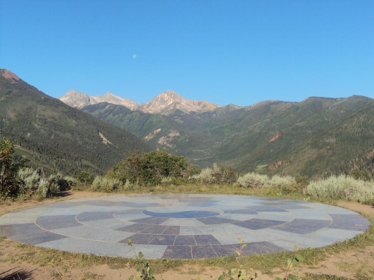 A scenic view of a circular stone platform surrounded by lush greenery, with majestic mountains in the background under a clear blue sky. A faint moon is visible in the sky above the mountains. The stone platform features a geometric design, creating a tranquil focal point in the natural landscape. Rim Trail mountain bike trail.