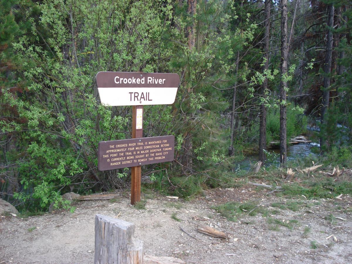 Sign marking the Crooked River Trail, surrounded by greenery and trees, with a stream visible in the background. The sign indicates that the trail is maintained for approximately four miles downstream and mentions that the trail is currently experiencing major disrepair, with efforts underway to address the issue. Crooked River mountain bike trail.