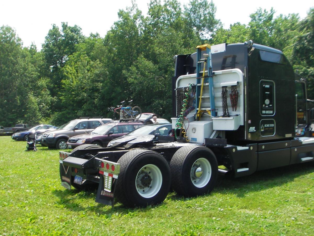 A black truck parked on grass, showing the rear view with a ladder resting against it. In the background, several cars are parked among trees in a green, outdoor setting. West Branch mountain bike trail.