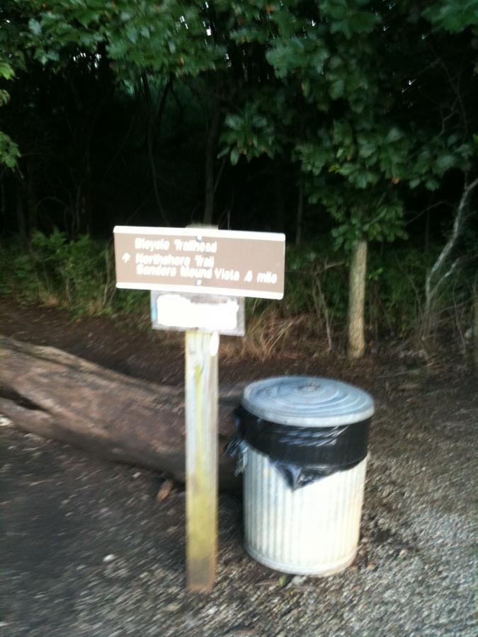 A brown wooden sign indicating a bicycle trailhead with directions to "Eagle's Nest Trail" and "Cedar's Scenic Vista 1 mile." Next to the sign is a metal trash can, partially obscured by a black bag, with greenery in the background. Clinton State Park mountain bike trail.