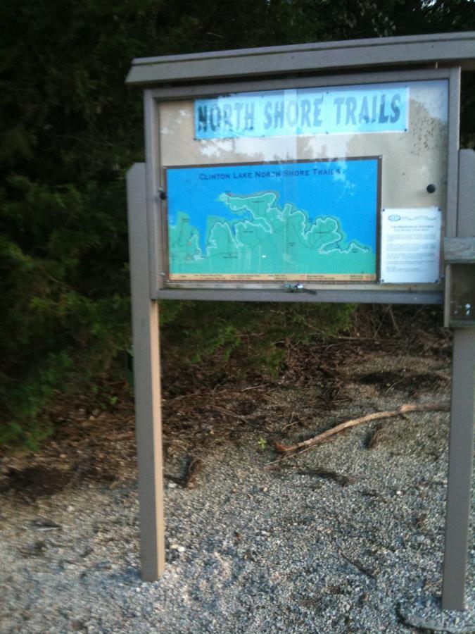 Information board detailing the North Shore Trails at Clinton Lake, featuring a map of the trails and instructions for visitors, surrounded by greenery and gravel. Clinton State Park mountain bike trail.