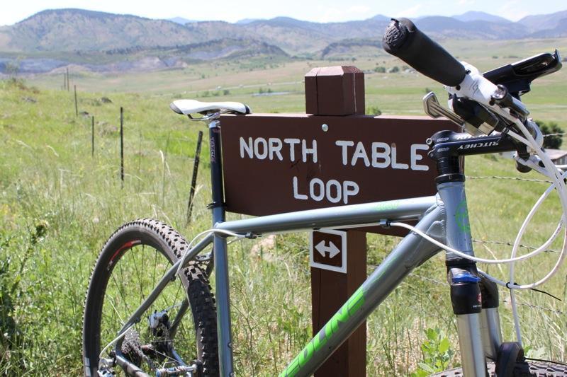 A mountain bike is parked next to a wooden sign that reads "North Table Loop," surrounded by green grass and rolling hills in the background under a clear blue sky. North Table Mountain mountain bike trail.