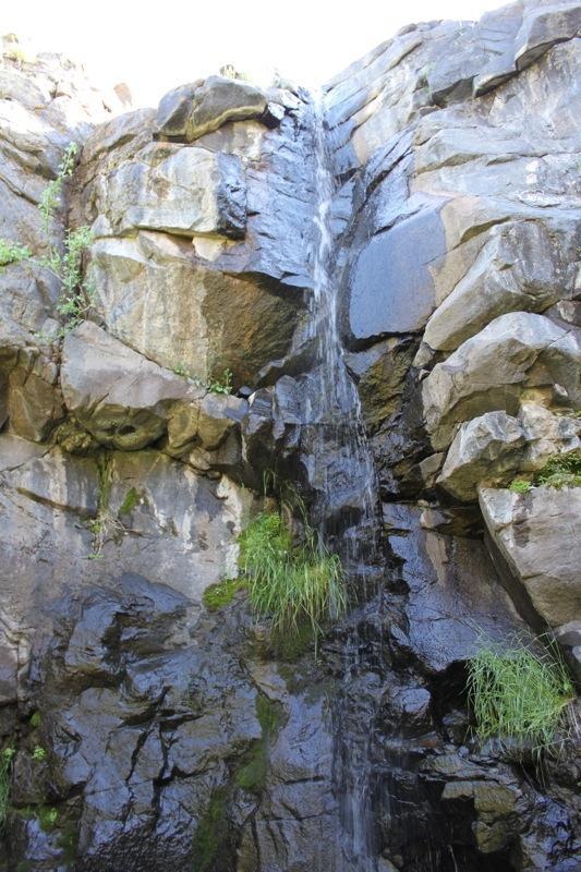 A small waterfall cascading down a rocky cliff, surrounded by dark stones and patches of greenery. The water flows gently over the rocks, creating a serene natural scene. Sunlight shines from above, illuminating the area. North Table Mountain mountain bike trail.