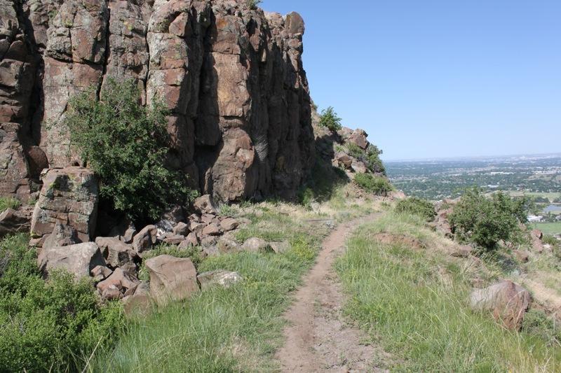 A rugged pathway winding along a rocky cliffside, surrounded by lush greenery and offering a view of the valley below under a clear blue sky. North Table Mountain mountain bike trail.