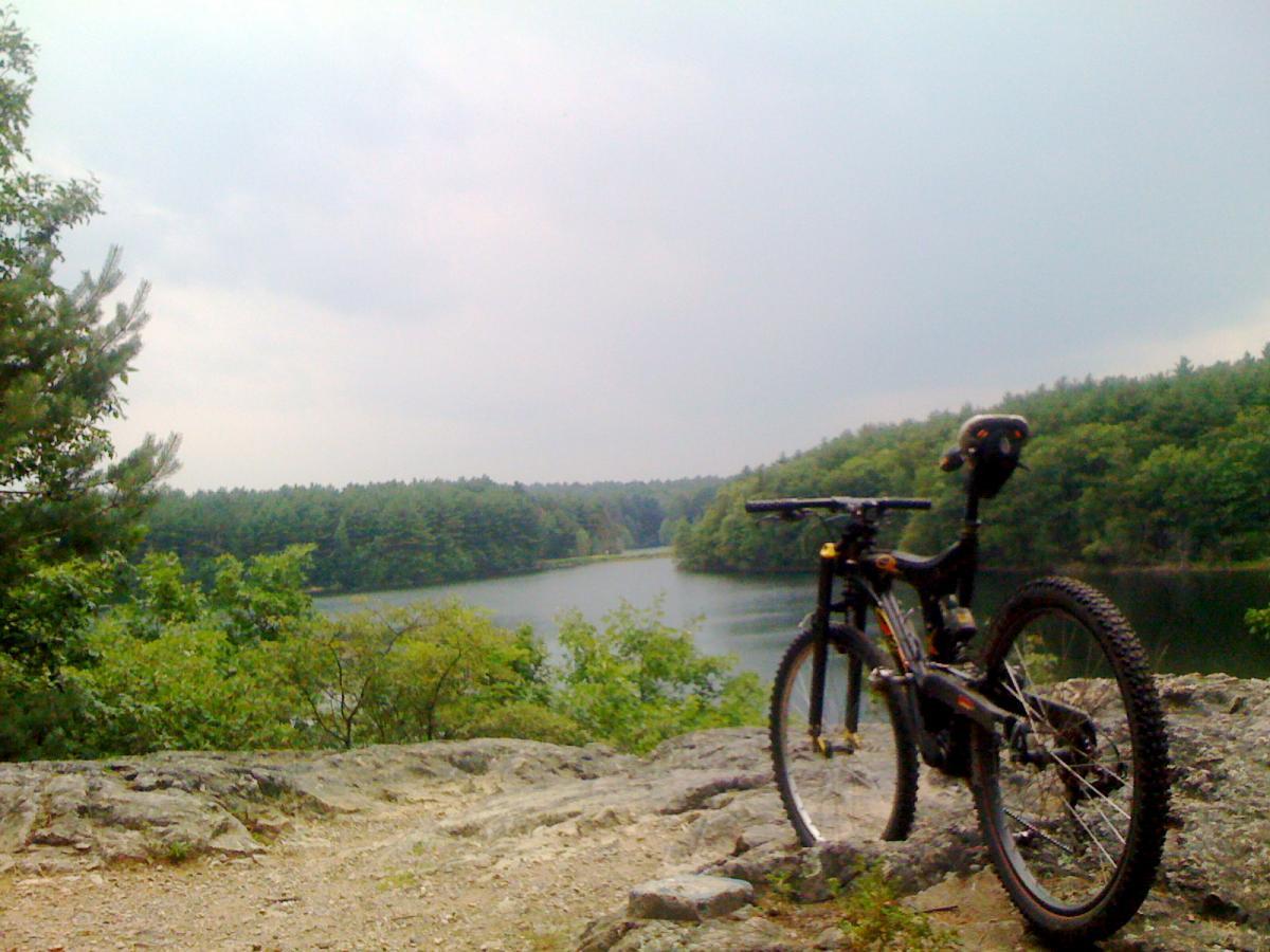 A mountain bike positioned on a rocky outcrop overlooking a calm lake surrounded by lush green trees under a cloudy sky. Middlesex Fells Reservation mountain bike trail.