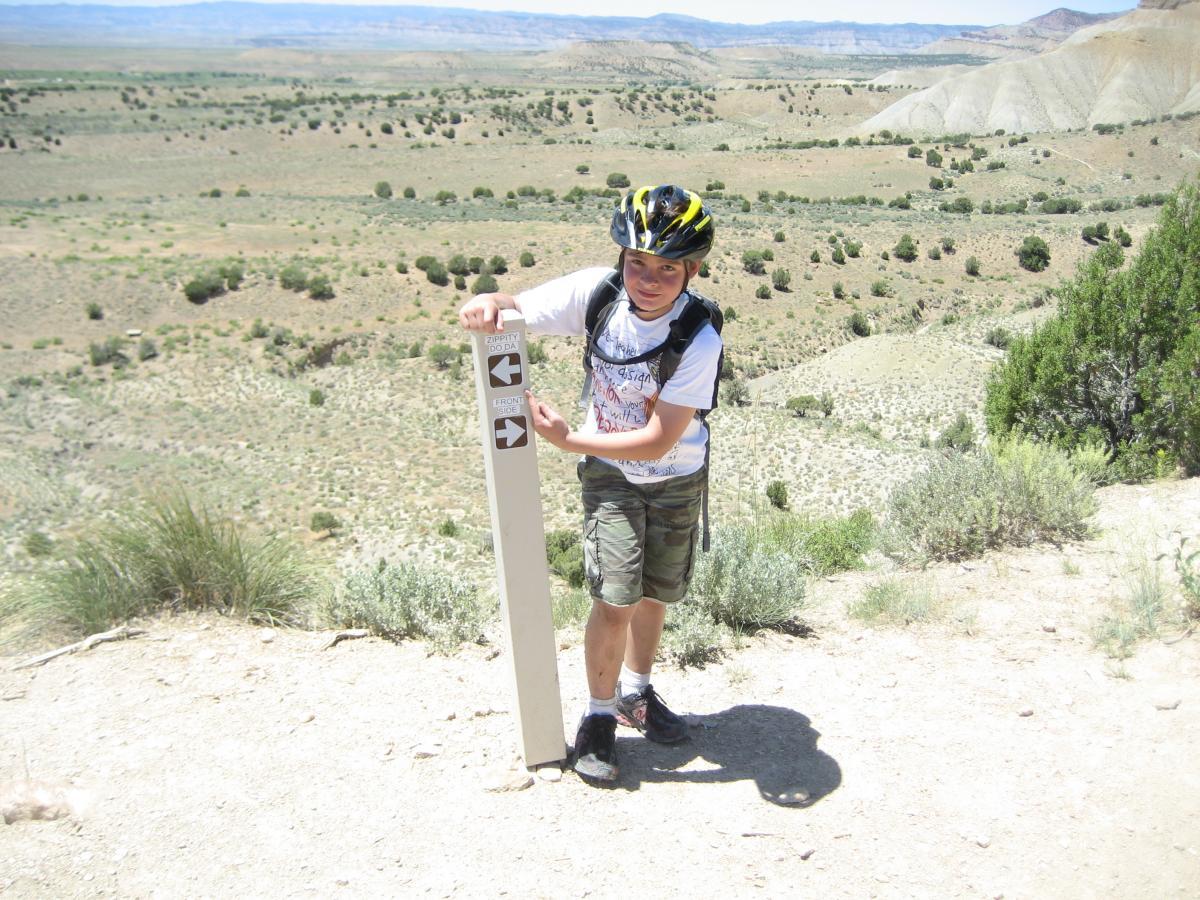 A child wearing a helmet and backpack stands beside a directional sign in a vast, arid landscape. The sign indicates paths to "Zipitti Zoo Dad" and "Eroded Slides." The background features a rolling terrain with sparse vegetation and distant hills under a clear blue sky. Zippety Do Dah mountain bike trail.