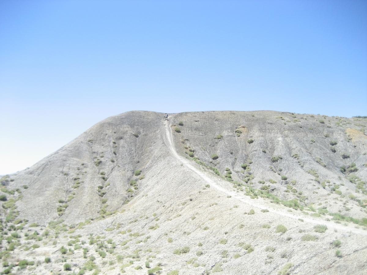 A dry, rugged hillside under a clear blue sky, featuring a winding dirt path leading to the top. Sparse vegetation, including small bushes, is scattered across the rocky terrain. Zippety Do Dah mountain bike trail.