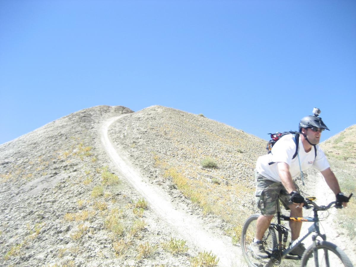 A mountain biker rides along a narrow dirt trail on a rocky hillside under a clear blue sky. The landscape is dotted with sparse vegetation, showcasing a rugged terrain. The biker is wearing a helmet equipped with a camera and casual riding attire, highlighting an adventurous outdoor activity. Zippety Do Dah mountain bike trail.