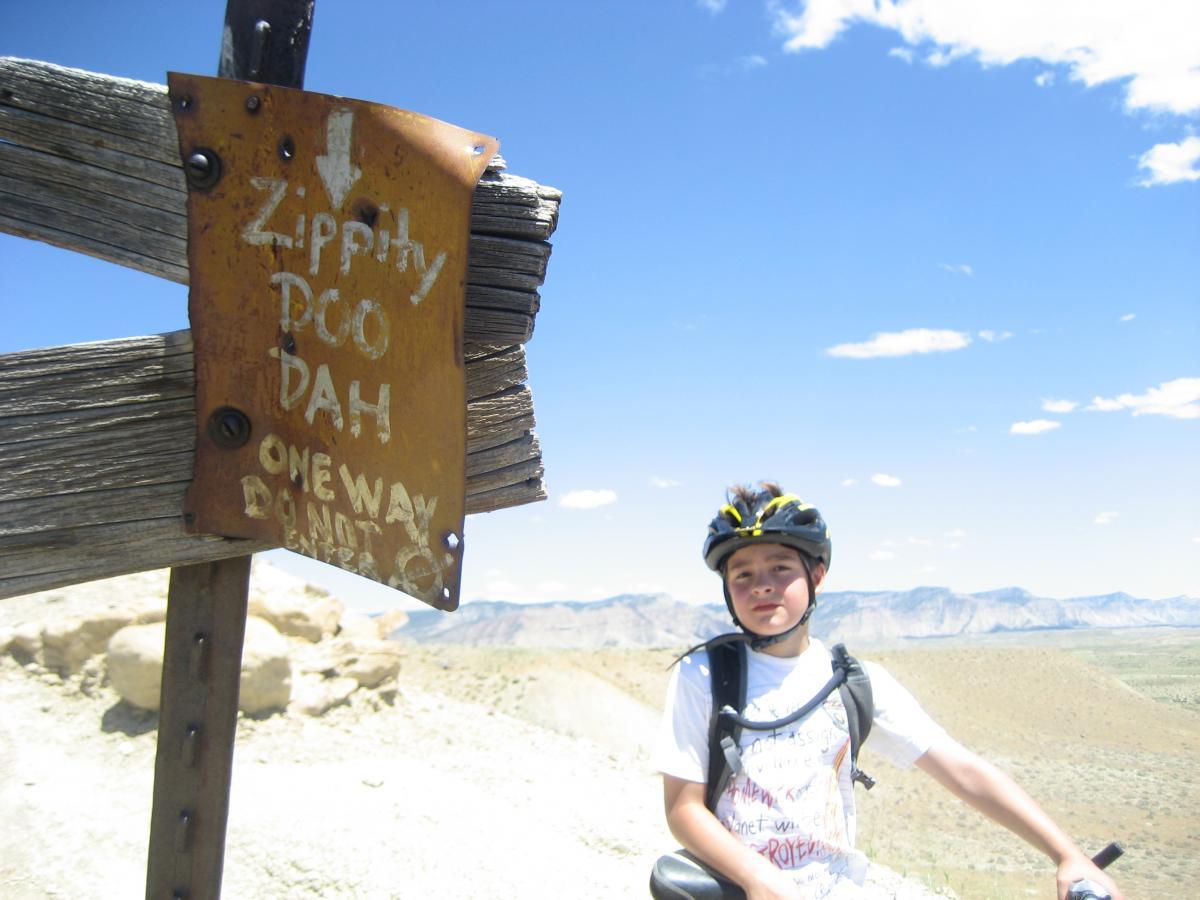 A young boy wearing a bicycle helmet and a t-shirt sits on a bike beside a wooden sign that reads "Zippity Doo Dah" with an arrow pointing down. The background features a clear blue sky and mountainous terrain in the distance. Zippety Do Dah mountain bike trail.