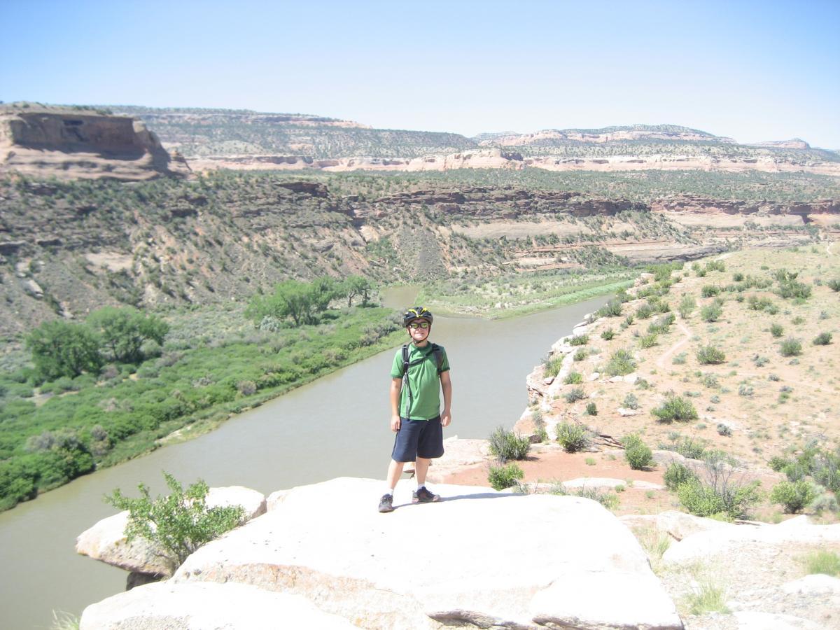 A young child wearing a green t-shirt and a helmet stands on a rocky ledge overlooking a winding river surrounded by green vegetation and desert landscapes under a clear blue sky. Mary's Loop / Horsethief Bench mountain bike trail.