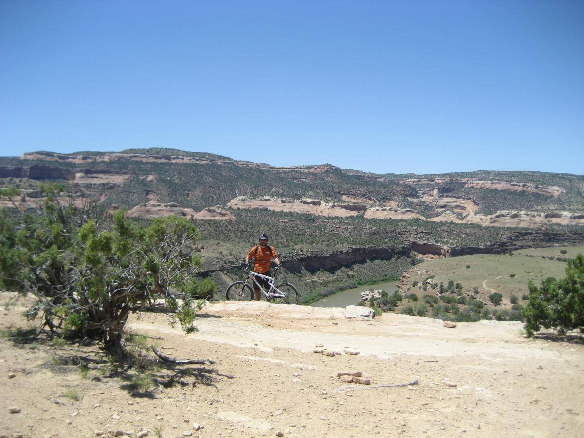 A mountain biker in an orange shirt stands beside their bike on a rocky trail, overlooking a sprawling canyon landscape with greenery and cliffs under a clear blue sky. Mary's Loop / Horsethief Bench mountain bike trail.