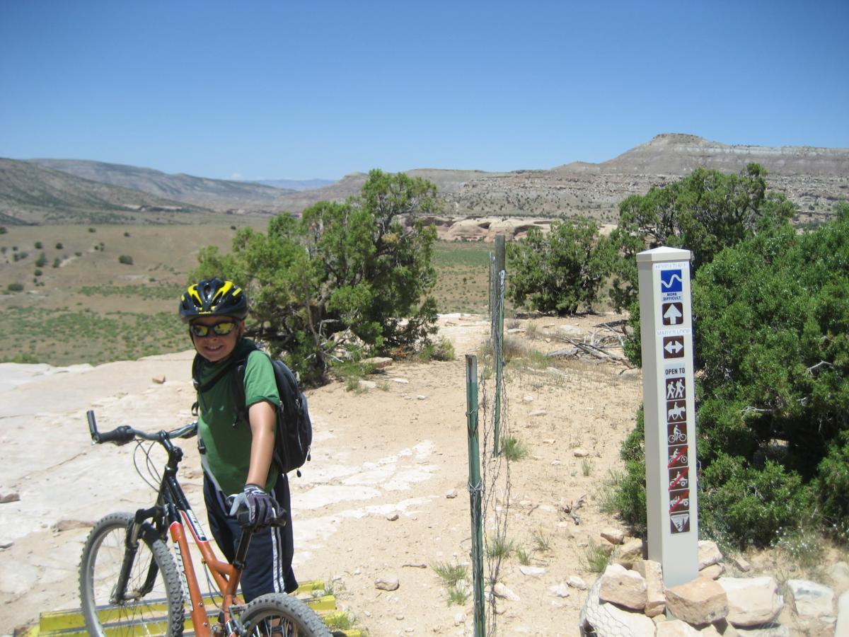 A young boy wearing a bicycle helmet and sunglasses smiles as he stands next to a mountain bike on a dirt trail. In the background, there are rolling hills and blue skies, and a trail sign indicating various outdoor activities. The area is surrounded by sparse vegetation typical of a rocky landscape. Mary's Loop / Horsethief Bench mountain bike trail.