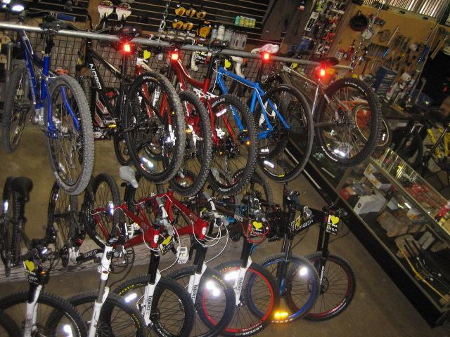 A variety of bicycles displayed in a shop, featuring several mountain bikes in different colors—blue, red, and white—hanging on a rack. Some bikes are equipped with lights on the rear, while others are positioned on the floor, showcasing their tires and handlebars. The background includes various bicycle accessories and equipment on display.