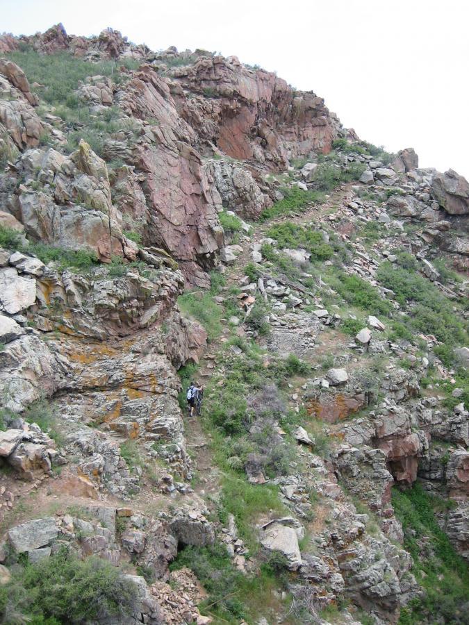 A person hiking along a rocky trail on a steep hillside, surrounded by greenery and boulders, under a cloudy sky. Curt Gowdy State Park mountain bike trail.