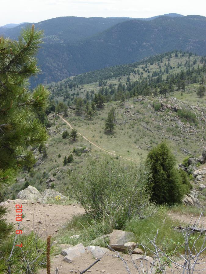 A panoramic view of a mountainous landscape, featuring rolling hills covered in greenery and rocky outcrops. A winding dirt path cuts through the scene, leading into the distance. Pine trees are scattered throughout the foreground and midground, adding to the natural beauty of the area under a partly cloudy sky. Centennial Cone Park mountain bike trail.