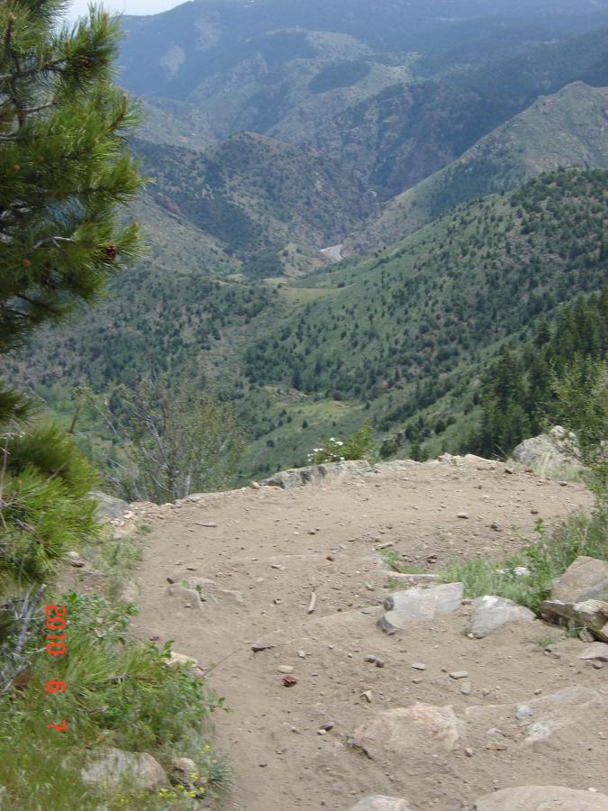 A scenic view of a mountainous landscape featuring rolling green hills and a valley below, with a winding dirt path leading into the distance. The foreground includes rocky terrain and patches of vegetation, while steep mountains rise in the background under a clear sky. Centennial Cone Park mountain bike trail.