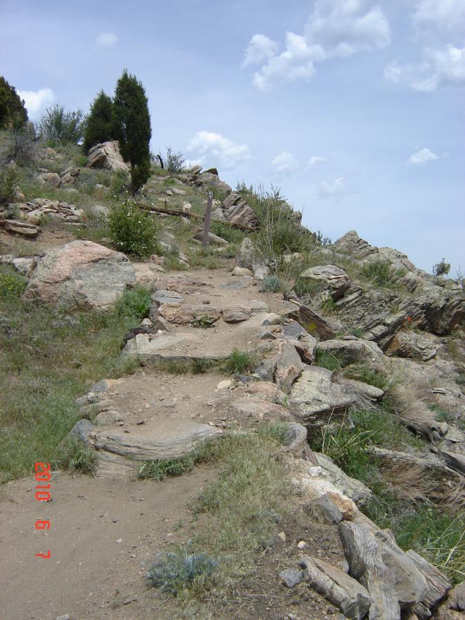 A winding dirt path leading upwards through rocky terrain, surrounded by sparse vegetation and small shrubs, under a cloudy blue sky. Centennial Cone Park mountain bike trail.
