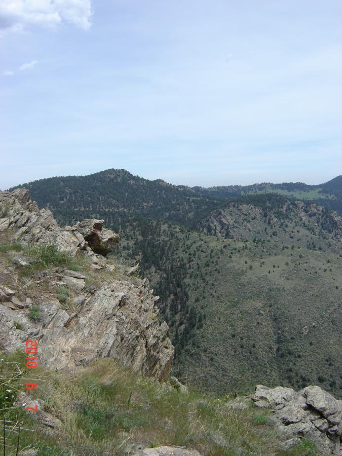 Scenic view from a rocky ledge overlooking a mountainous landscape, with green hills and trees in the distance under a partly cloudy sky. Centennial Cone Park mountain bike trail.