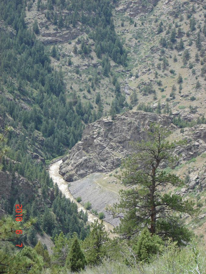 A scenic view of a river winding through a mountainous landscape, surrounded by dense green forests and rocky cliffs. The river's water appears muddy, indicating recent rainfall, while trees dot the slopes of the hills. The image captures the natural beauty of an outdoor setting. Centennial Cone Park mountain bike trail.