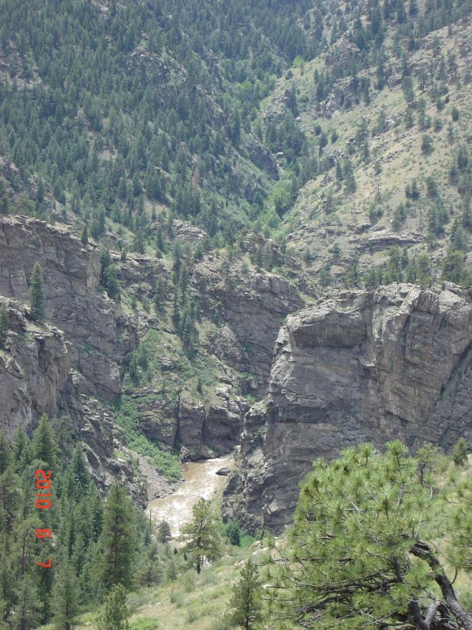 A scenic view of a rocky canyon surrounded by dense greenery, with a river flowing through the bottom of the gorge. The landscape features steep cliffs and a variety of trees, highlighting the natural beauty of the area. Centennial Cone Park mountain bike trail.