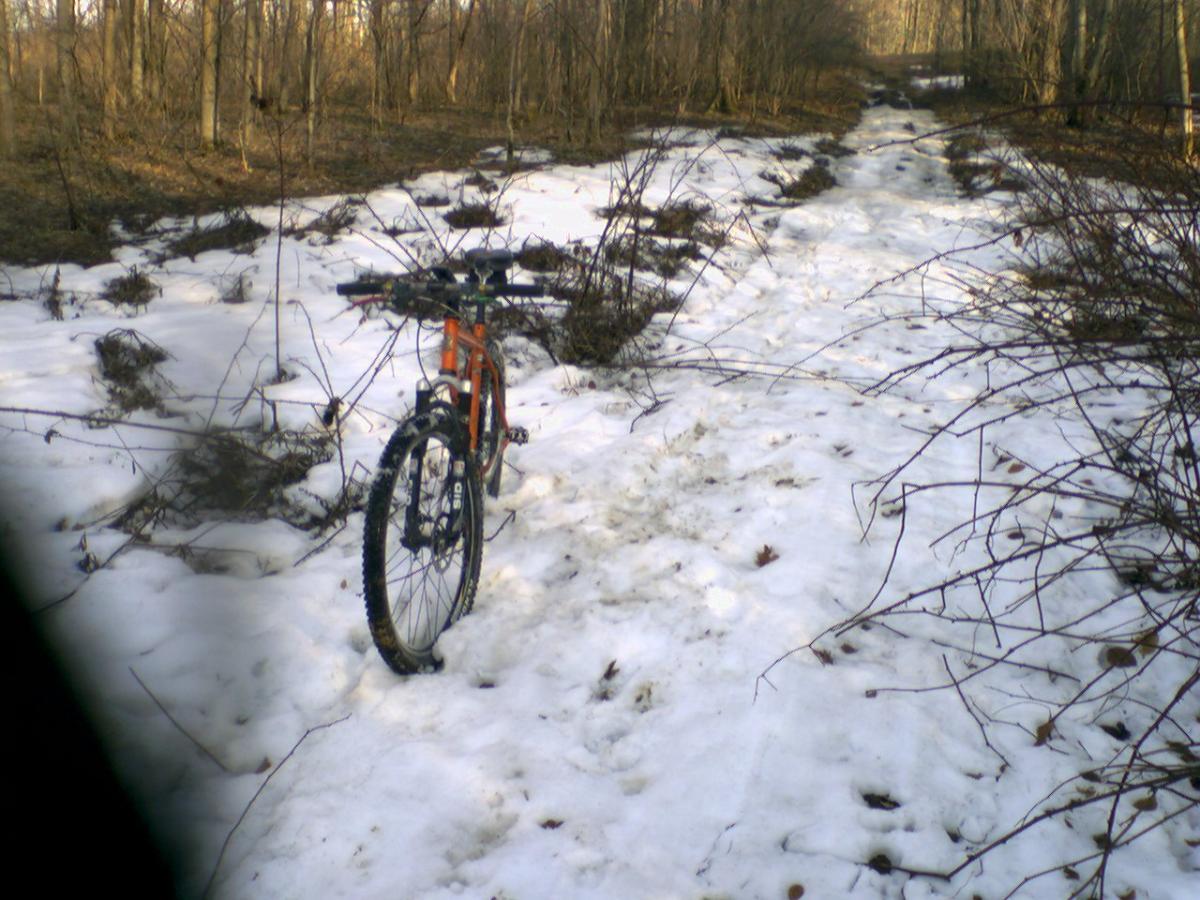 A mountain bike rests on a snowy, uneven trail surrounded by trees. The path is partially covered in snow, with tire tracks leading into the distance, and tangled branches are visible along the sides of the trail. The scene conveys a quiet, winter landscape ideal for off-road cycling. Sherrillbrook Park mountain bike trail.