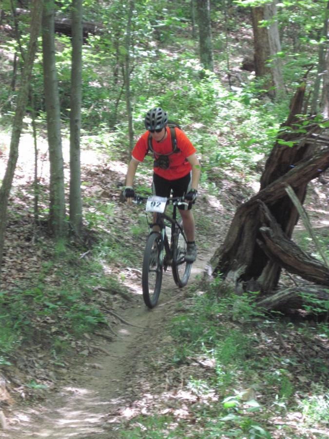 A person riding a mountain bike on a narrow trail through a densely wooded area, wearing a helmet and an orange shirt, with a race number visible on their bike. Sunlight filters through the trees, illuminating the greenery and leaf-covered ground. Raccoon Mountain Trail Network mountain bike trail.