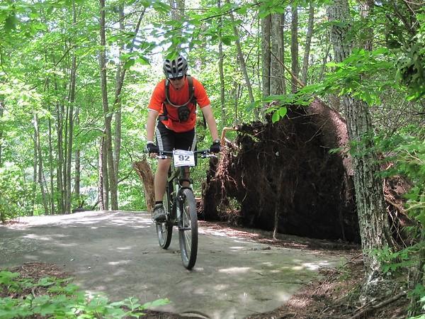 A mountain biker in an orange shirt and helmet rides along a forested trail, with lush green trees surrounding them. In the background, an uprooted tree stump is visible, emphasizing the natural setting of the outdoor scene. Raccoon Mountain Trail Network mountain bike trail.