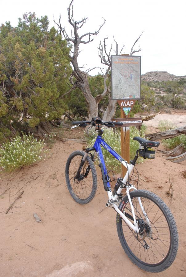 A mountain bike stands next to a trail map sign in a desert landscape, surrounded by green shrubs and dry soil, with distant hills in the background. Klondike Bluffs mountain bike trail.