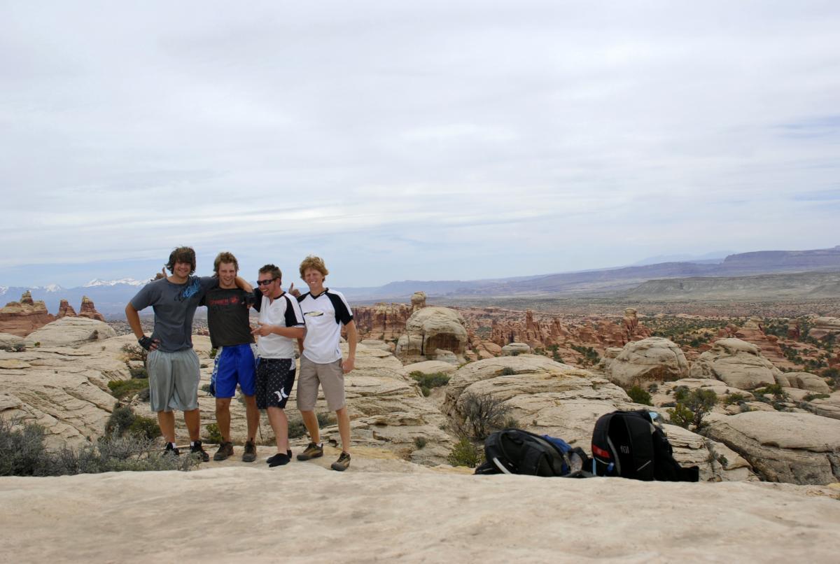 Four young men stand together on rocky terrain, smiling and posing for the camera. They are dressed in casual outdoor attire, with backpacks resting on the ground beside them. The landscape in the background features unique rock formations and a vast expanse of desert, under a cloudy sky. Klondike Bluffs mountain bike trail.