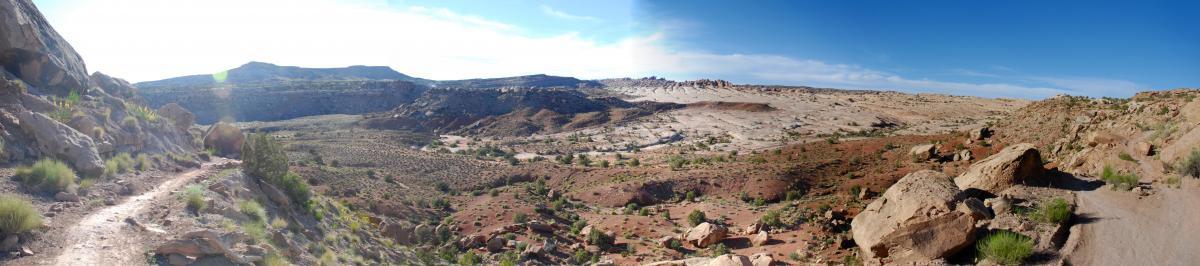 A panoramic view of a rugged landscape featuring rocky terrain, scattered vegetation, and distant hills under a clear blue sky. A dirt path winds through the foreground, leading into the expansive valley and rolling hills beyond. Sovereign Single Track mountain bike trail.