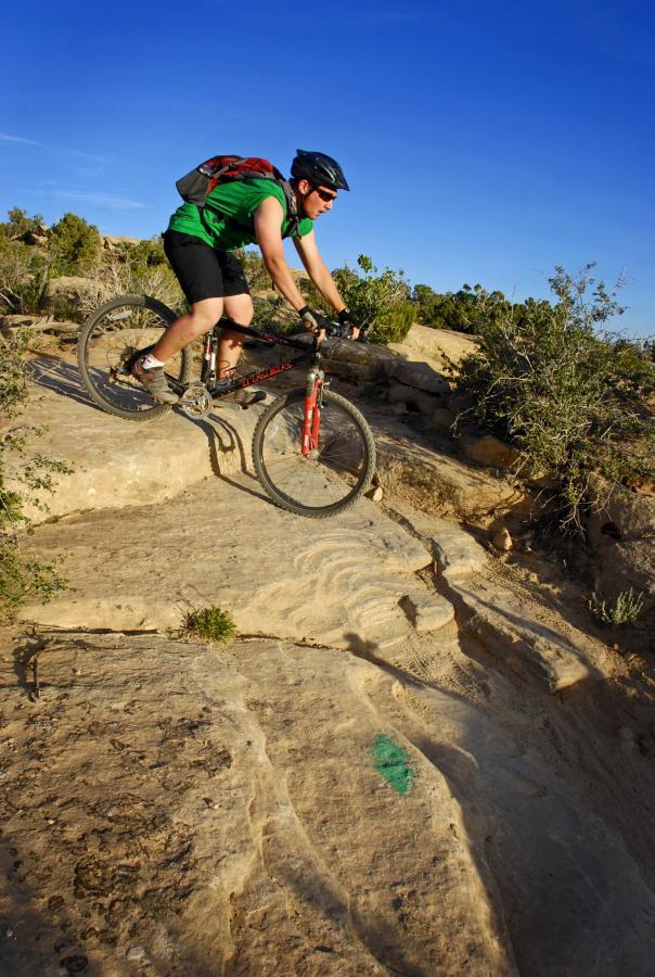 A mountain biker wearing a green shirt and a backpack navigates a rocky trail under a clear blue sky. The bike is positioned on rugged terrain with visible markings on the rocks, surrounded by sparse vegetation. Sovereign Single Track mountain bike trail.