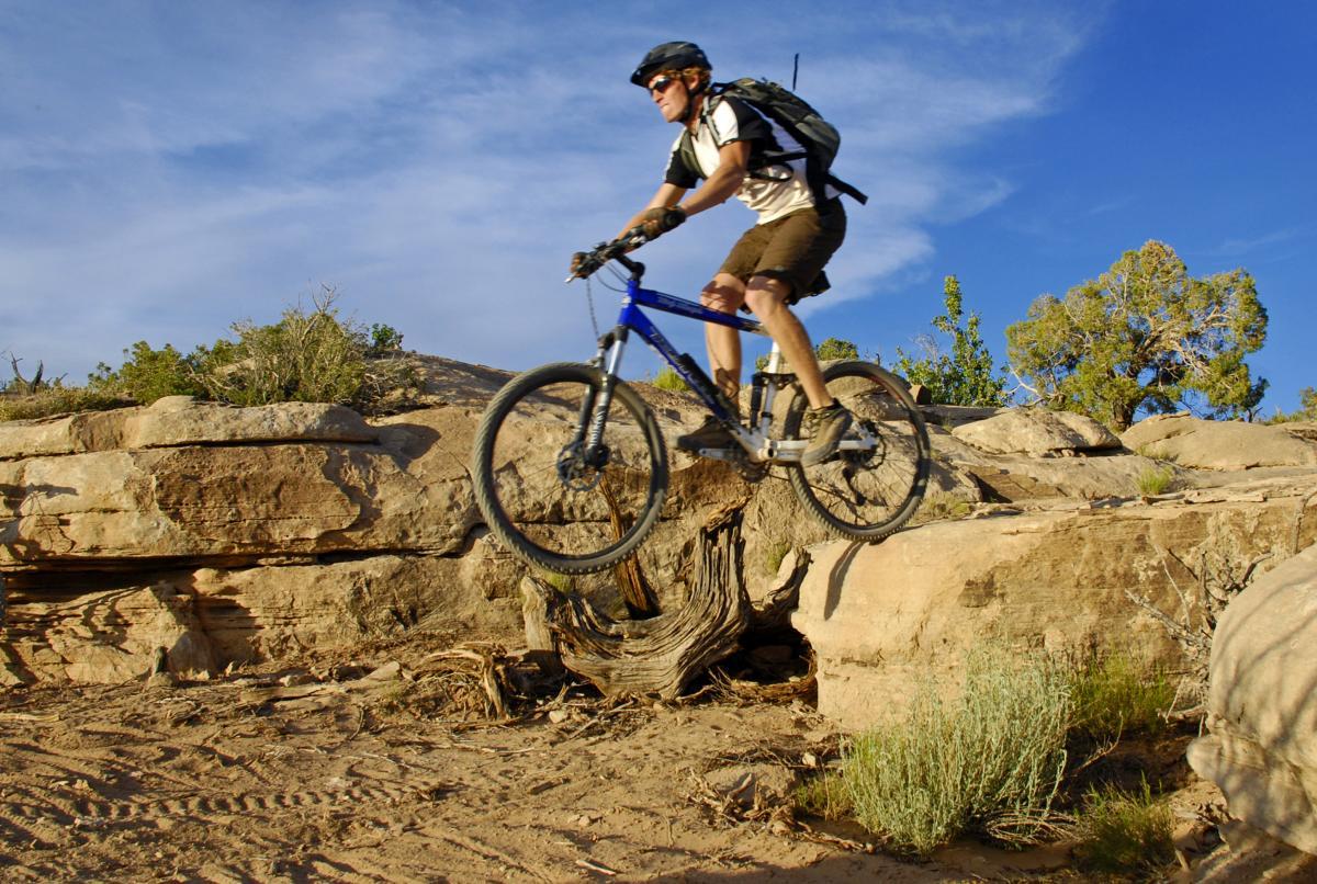 A mountain biker jumping off a rocky ledge, with a blue mountain bike and wearing a helmet and sunglasses. The background features a rugged landscape with shrubs and trees under a clear blue sky. Sovereign Single Track mountain bike trail.